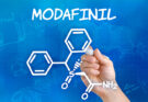 Hand holding a pen drawing the chemical formula of Modafinil on a sheet of paper. The chemical structure is composed of various shapes and lines, including hexagons, triangles, and circles, with letters and numbers interspersed throughout.