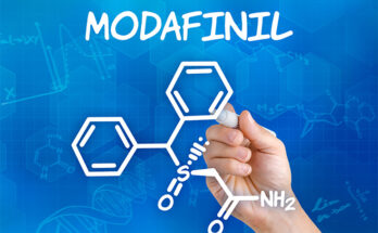 Hand holding a pen drawing the chemical formula of Modafinil on a sheet of paper. The chemical structure is composed of various shapes and lines, including hexagons, triangles, and circles, with letters and numbers interspersed throughout.